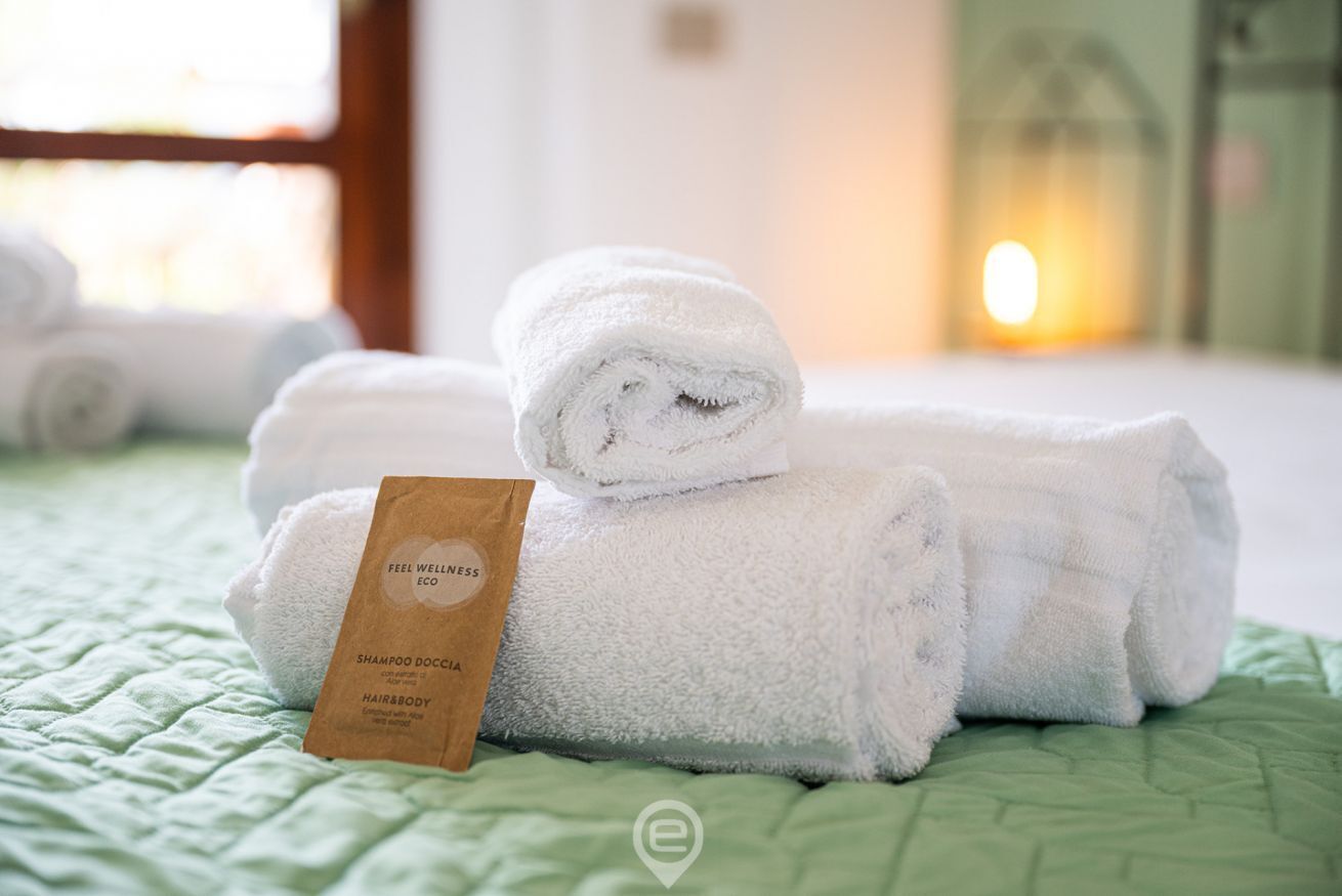 En pænt redt seng med sammenrullede hvide håndklæder og en brun papirpose med påskriften "FEEL WELLNESS ECO SHAMPOO DOCCIA HAIR&BODY".