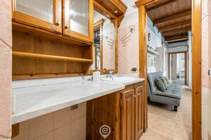 Ein Badezimmer mit Holzausstattung, einem Waschbecken und einem Spiegel. Im angrenzenden Wohnbereich steht ein graues Sofa.