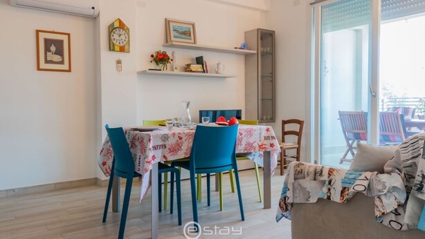 Zona pranzo con tavolo imbandito per quattro persone, sedie, orologio e mensole con oggetti decorativi.