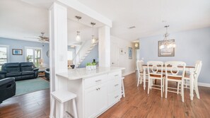 A modern kitchen with white cabinetry, a dining area with wooden chairs, and a staircase leading to another level. - Amazing Beachside Pool Home 5 Bedrooms - Summer Days Cooler NIghts (Wilbur by the Sea)
