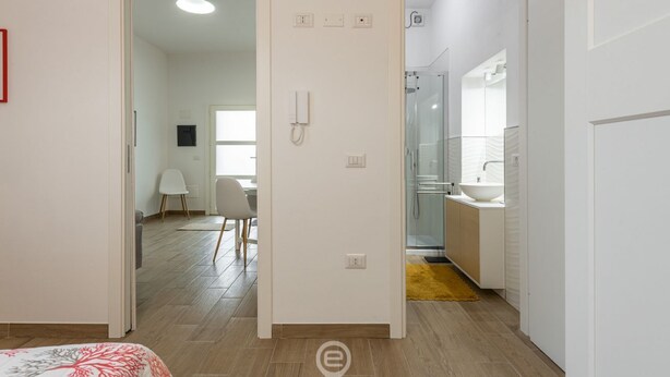 A modern hotel room with a wooden floor, a bed with a floral bedspread, a bathroom with a glass shower, and a small sitting area with a chair and table.