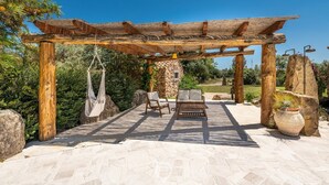 An outdoor patio with a wooden pergola, stone pillars, a hammock, and a seating area with a table and chairs.