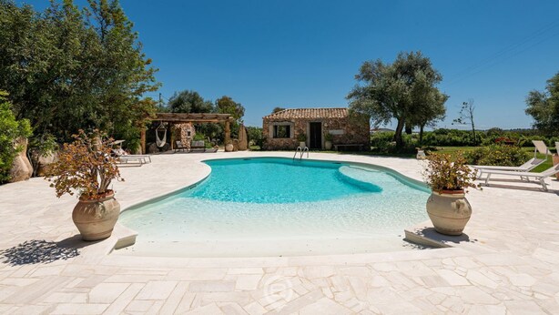 A swimming pool with a stone deck, surrounded by trees and a small stone building.