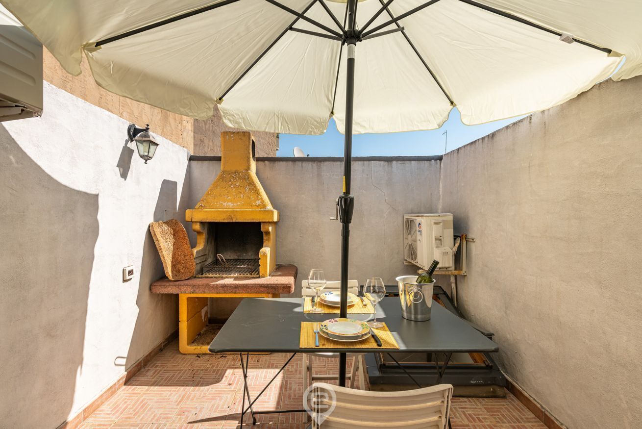 An outdoor patio with a table set for dining, a grill, and a large umbrella providing shade.