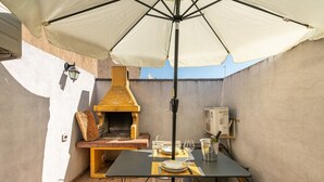 An outdoor patio with a table set for dining, a grill, and a large umbrella providing shade. - Maty's House (Gonnesa)