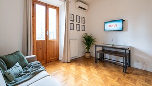 A living room with a sofa, a wooden floor, a table, a potted plant, a wall-mounted TV displaying 'NETFLIX', and a window with curtains. - Colosseum ★★★★★ - Rooftop with HOT jacuzzi and Skyline of Rome (Roma)