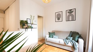 Una sala de estar moderna con un sofá blanco, cojines verdes y piso de madera. Hay obras de arte enmarcadas en la pared y una planta en la esquina.