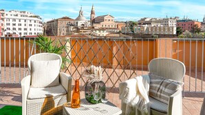 Un balcón con sillas de mimbre, una mesa con una botella y vasos, y vistas a edificios históricos.