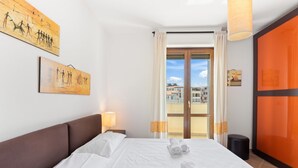 A hotel room with a large bed, two paintings on the wall, a window with curtains, and a view of buildings outside. - Antonella's Apartment (Alghero)