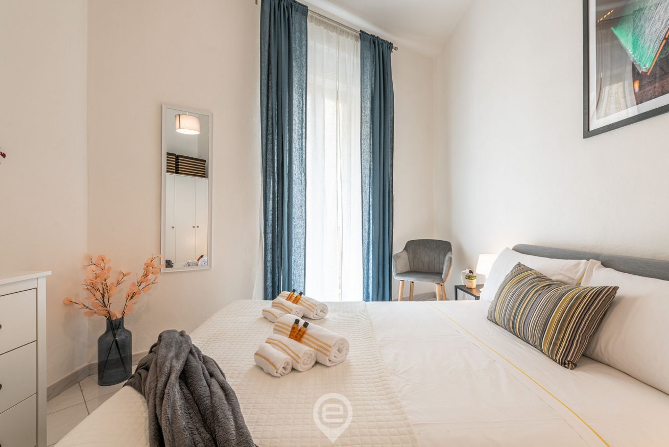 A modern hotel room with a large bed, a bedside table, a chair, a mirror, and a window with curtains.