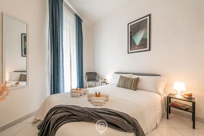 A hotel room with a large bed, a bedside table with a lamp, a chair, a mirror, and a painting on the wall. - Aurora’s Apartment: the perfect retreat in the heart of Cagliari (Cagliari)