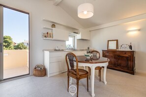 A modern kitchen with white cabinetry, a dining table with chairs, a hanging light fixture, and a view of the outdoors.