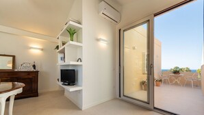 Una sala de estar moderna con una televisión, estanterías blancas y un ventanal corredera que da a un balcón con mobiliario exterior.