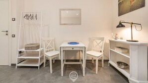 A room with a white desk, two white chairs, a small table with a blue tray, a white shelf with a lamp, and a painting on the wall.