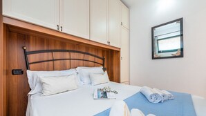 Ein ordentlich gemachtes Bett mit weißer Bettwäsche, einem blauen Akzent und aufgerollten Handtüchern. Auf dem Bett liegt eine Zeitschrift, und im Hintergrund ist ein kleines Fenster mit Vorhängen zu sehen.