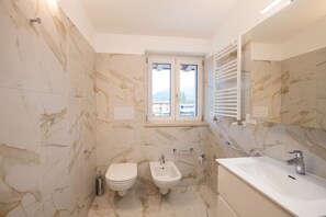 Een badkamer met toilet, bidet en bad, marmeren wanden en een raam met uitzicht.