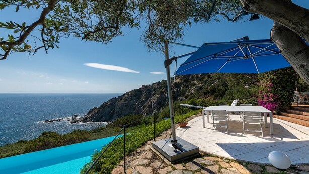 En terrasse med et bord og stole, en blå parasol og havudsigt.