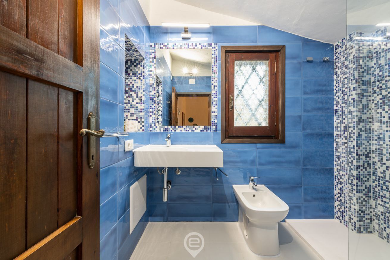 Uma casa de banho com azulejos azuis, uma porta de madeira, uma pia, um bidé e um espelho.