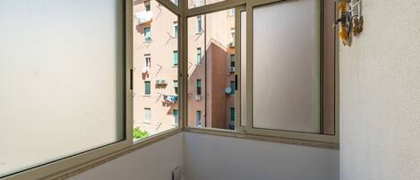 A balcony with a view of neighboring buildings, a window with a view, and a wall-mounted air conditioning unit.
