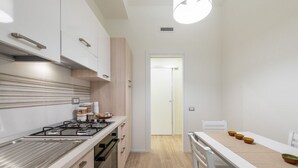 Una cuina moderna amb fogó a gas, forn i armaris blancs. Hi ha una zona de dinar amb taula i cadira, i una porta que dona a una altra habitació.