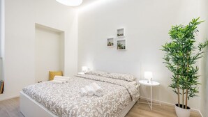 Un dormitori modern amb un llit gran, taules de nit, una planta en test i una petita estant amb objectes decoratius.