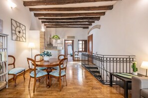 Un coin repas avec des chaises et une table en bois, un escalier avec une rampe en fer forgé, et une cuisine en arrière-plan.