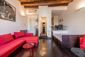 Un salon moderne avec un canapé rouge, une petite table ronde et un lit doté d’une tête de lit en bois. Un escalier mène à un espace cuisine.