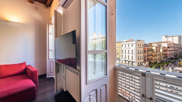 A balcony with a red sofa, a TV, and a city view. - Piazza Yenne Apartment (Cagliari)