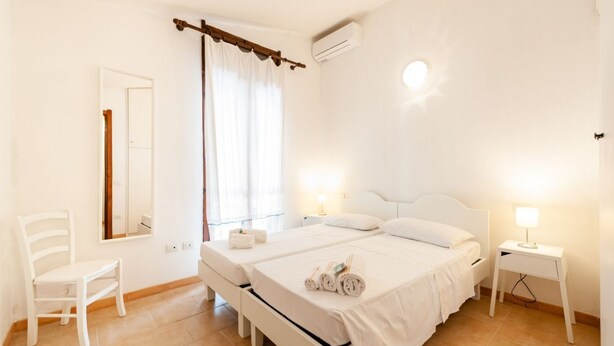 Een schone, witte kamer met twee bedden, een stoel, een spiegel en een raam met gordijnen.