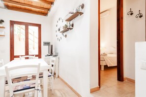 A room with a white table and chairs, a bed with a white duvet, and a wall-mounted shelf with decorative items.
