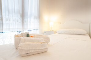 Habitación de hotel con toallas ordenadamente dobladas sobre la cama, una lámpara de noche y una ventana con cortinas traslúcidas.