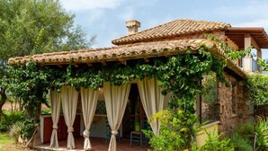 A rustic stone house with a tiled roof, a covered porch with curtains, and a lush garden.