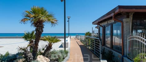 A coastal walkway with a restaurant named "La Cantina del Mar" on the right, palm trees, and a clear view of the sea.