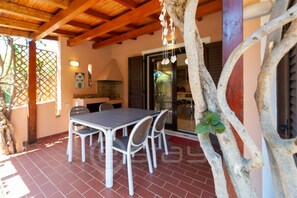 Um alpendre coberto com mesa e cadeiras, teto de madeira e vista do interior através de uma porta de vidro.