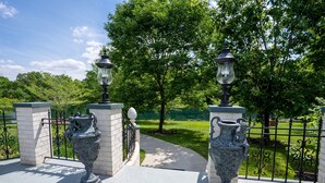 Zwei verzierte Urnen mit Lampen auf Sockeln, ein gepflasterter Weg und ein Metallzaun mit Blick auf einen grünen Park.