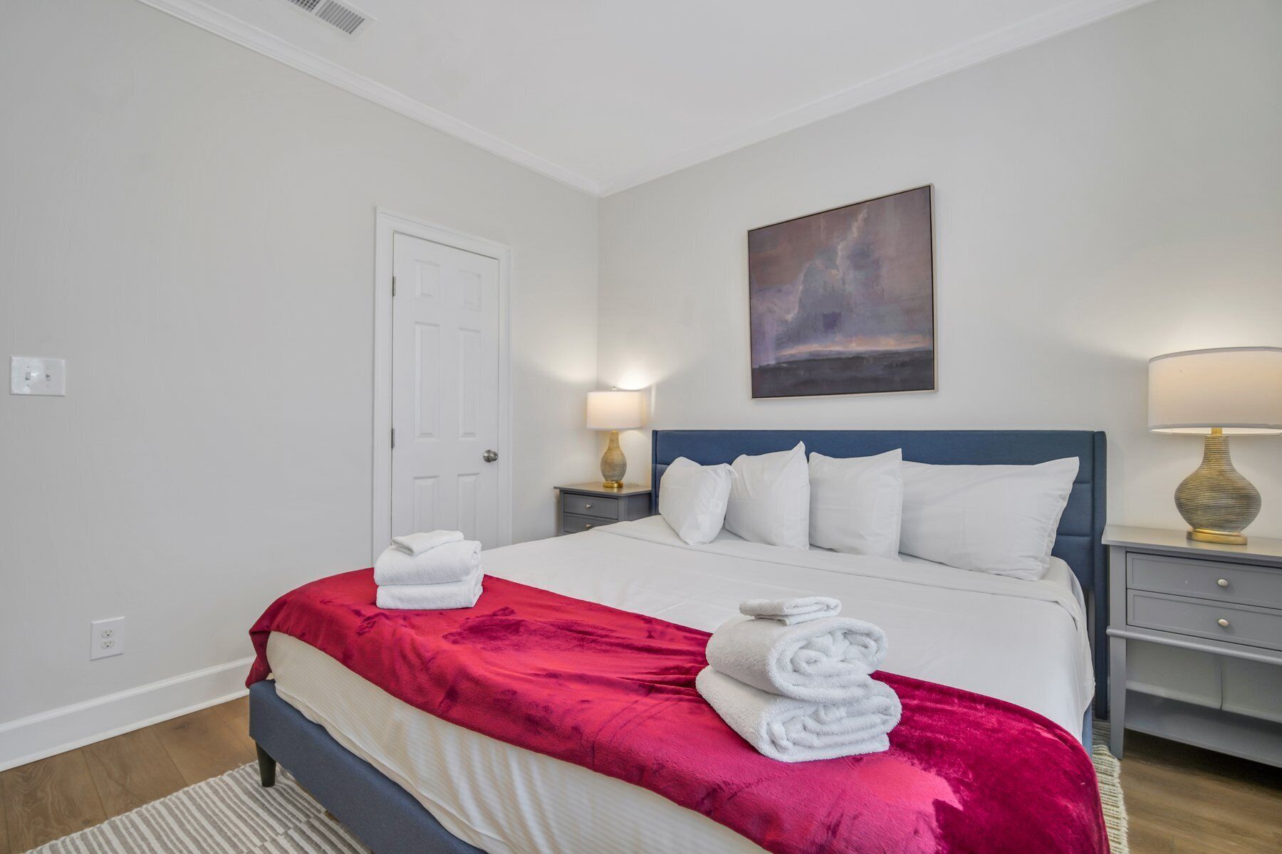 A hotel room with a bed, two bedside lamps, a painting on the wall, and a door.