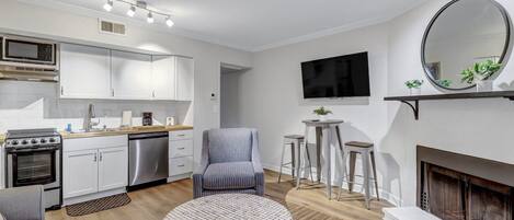 Eine moderne Küche mit weißen Schränken, einer eingebauten Mikrowelle und einem Edelstahl-Geschirrspüler. Es gibt ein graues Sofa, einen runden Couchtisch und einen Kamin mit einem Spiegel darüber.