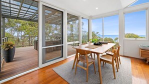 Un comedor moderno con una mesa y sillas de madera, ventanas amplias que ofrecen vista al mar y un balcón con una planta en maceta.