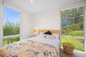 A bedroom with a large bed, a nightstand, a lamp, and a view of a garden through a large window. - Big Hill Retreat (Lorne)