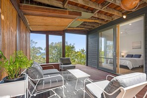A modern outdoor seating area with a view of the sea, a glass door leading to a bedroom, and a wooden ceiling.