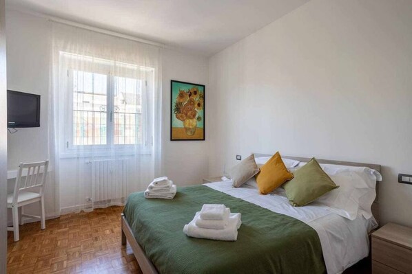 Un dormitorio con cama, almohadas, una silla, un cuadro y un televisor.
