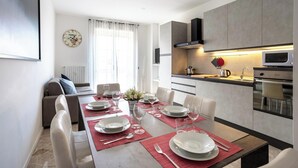 Un comedor moderno con una mesa dispuesta para cuatro, una cocina con microondas y estufa, y una sala de estar con sofá y un reloj en la pared.