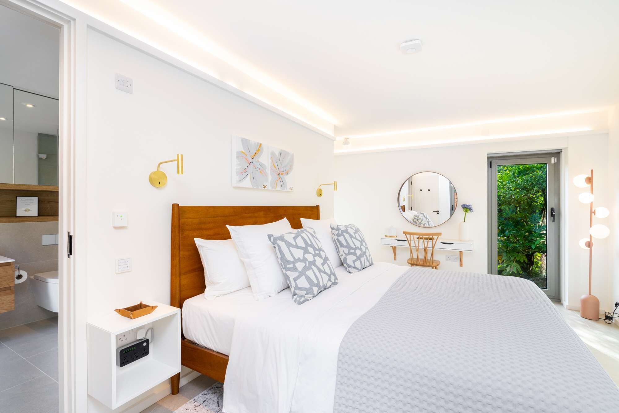 A modern bedroom with a wooden bed, bedside tables, a vanity with a mirror, and a view of greenery outside.