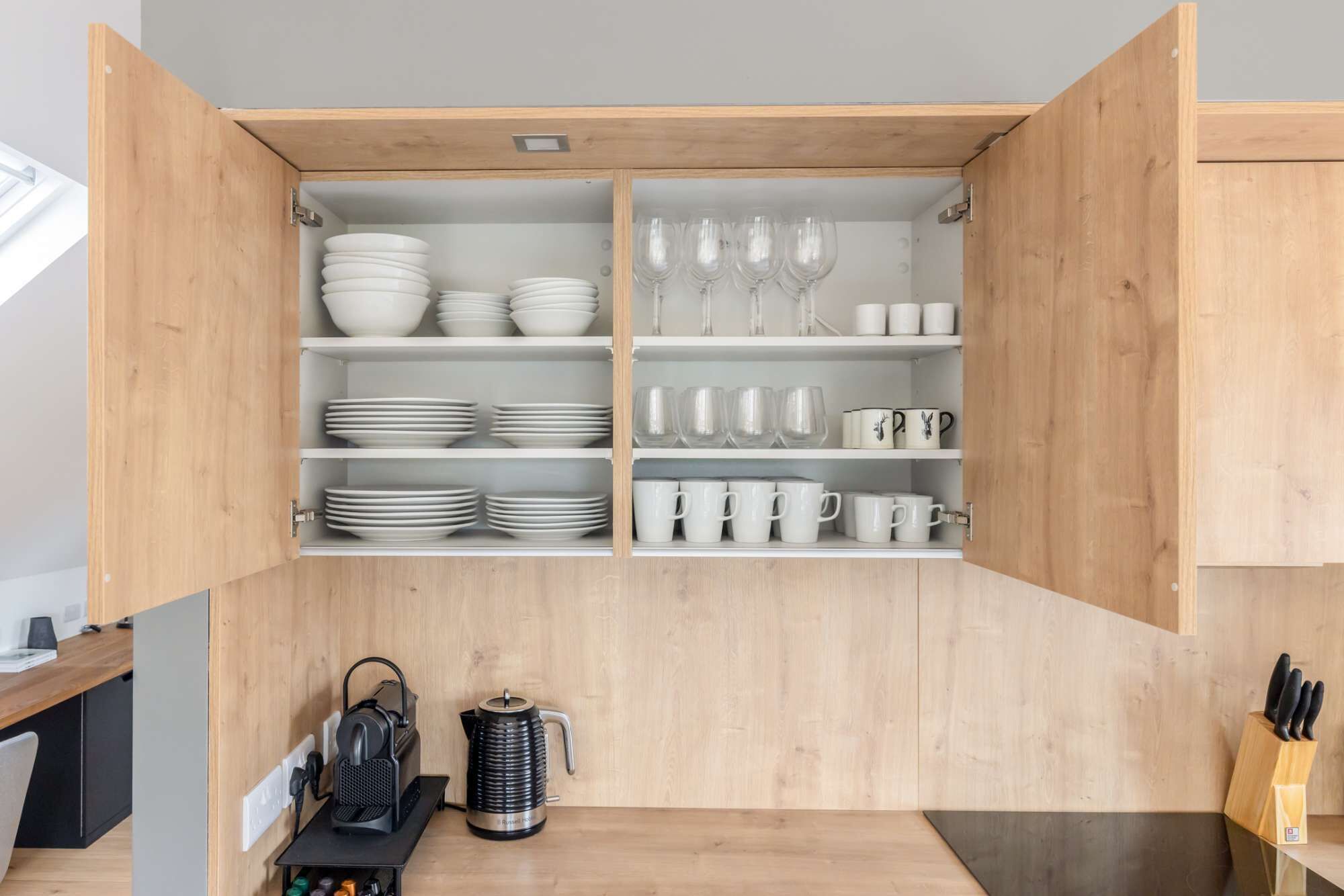 A kitchen with wooden cabinets, shelves with white dishes and glasses, a coffee maker, and a knife block.