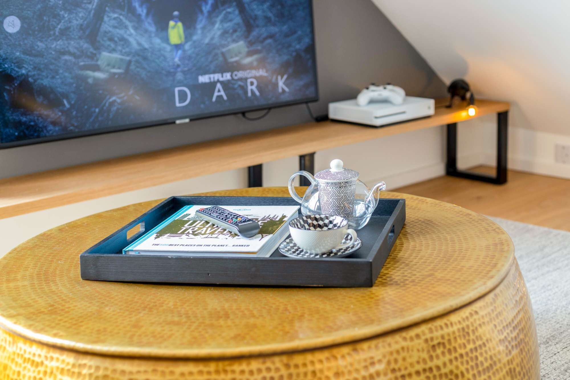 En hyggelig stue med et fjernsyn der viser 'Netflix Original Dark', et sofabord med en bakke med en bog og en tekedel, og en spillekonsol på en hylde.