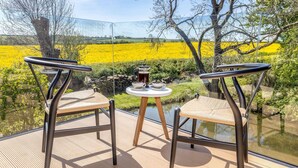 Una terraza con dos sillas de mimbre y una mesita pequeña, con vistas a un campo de flores amarillas y una masa de agua.