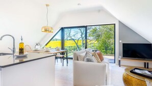 Un loft moderno con cocina, comedor y sala de estar. Cuenta con un amplio ventanal con vista a un campo amarillo, un televisor de pantalla plana y una mesa de comedor preparada para cuatro personas.