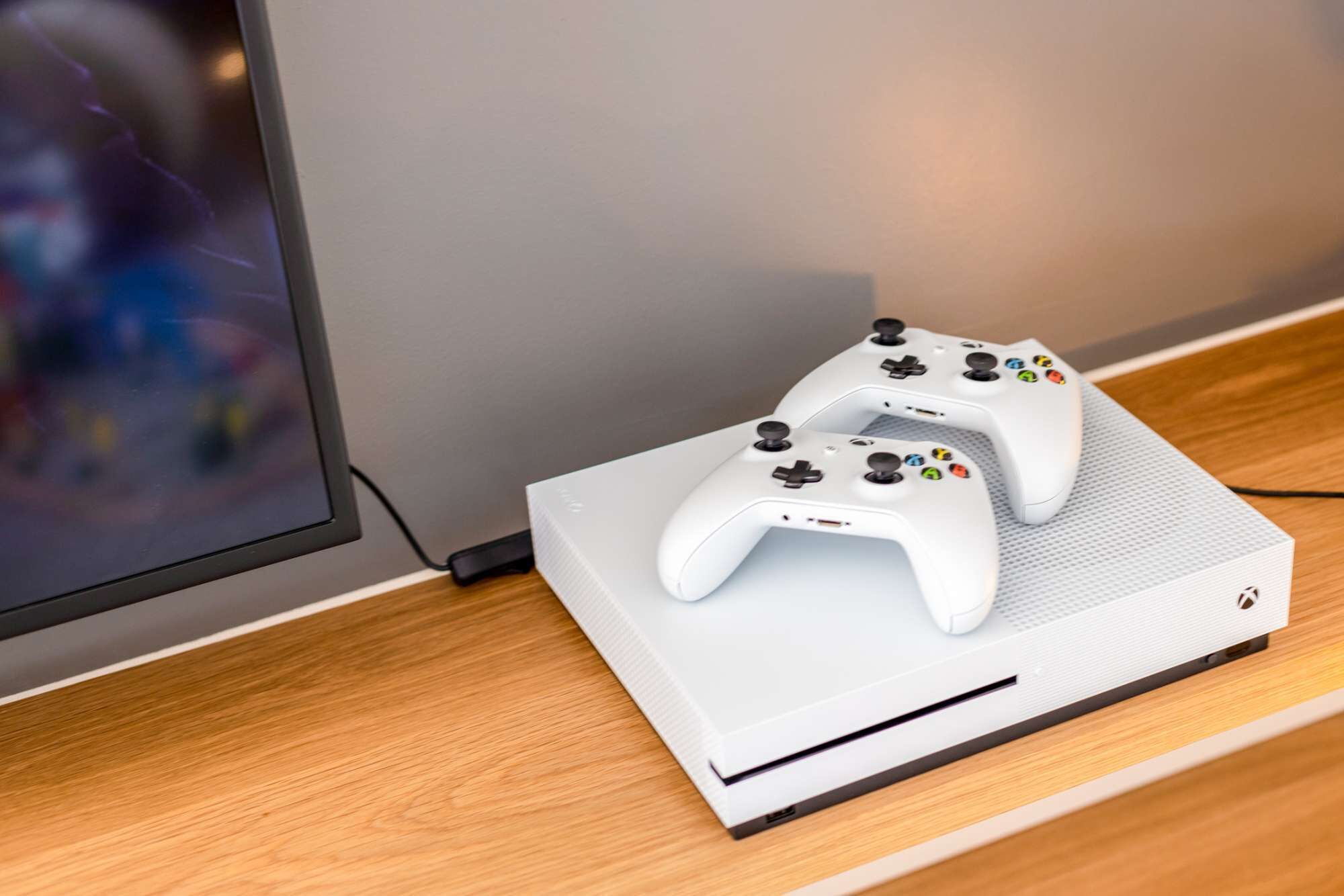 A white Xbox console with two controllers on a wooden surface next to a monitor.