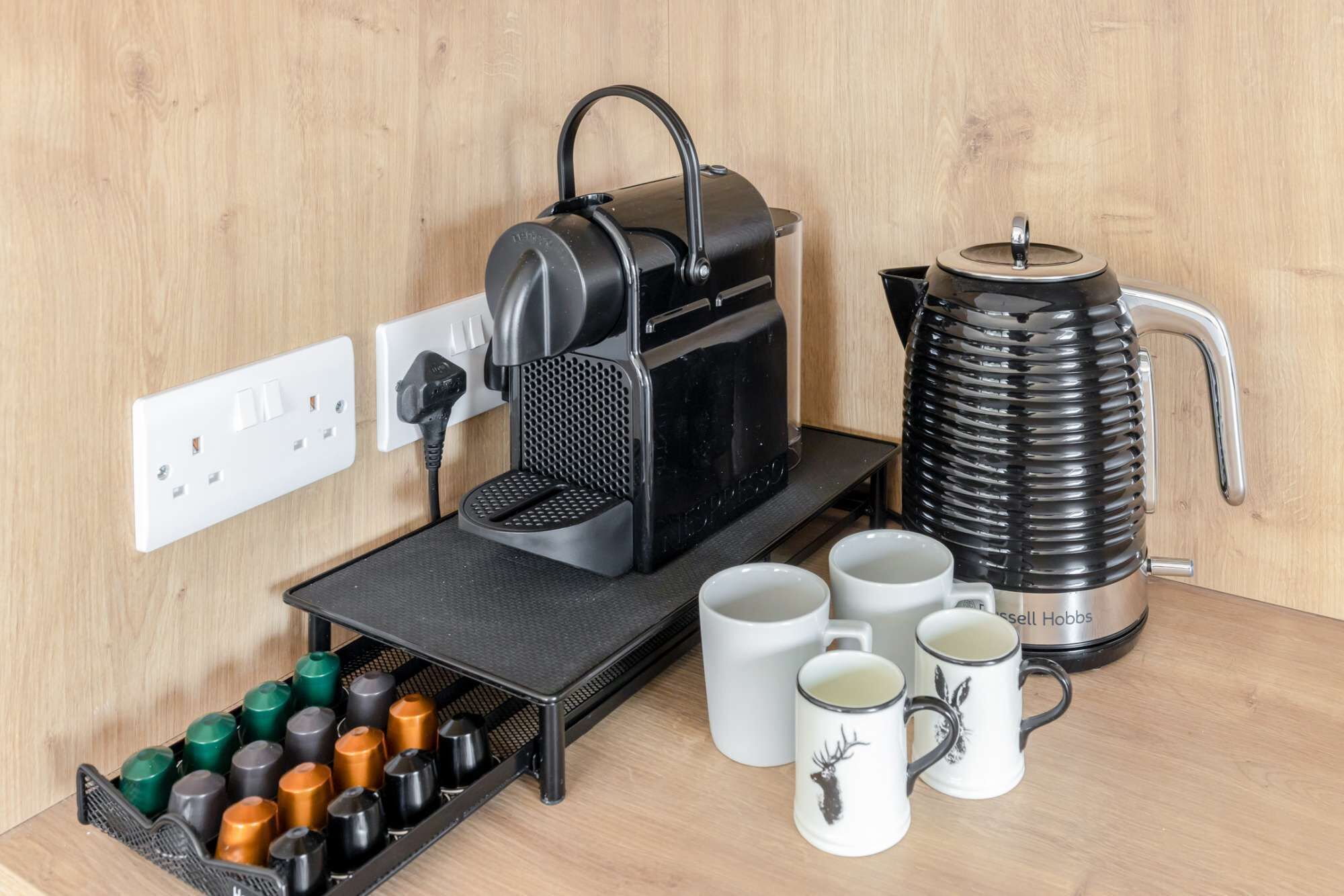 Eine Kaffeemaschine, ein Wasserkocher und ein Becherhalter mit mehreren Tassen auf einem Holztisch.