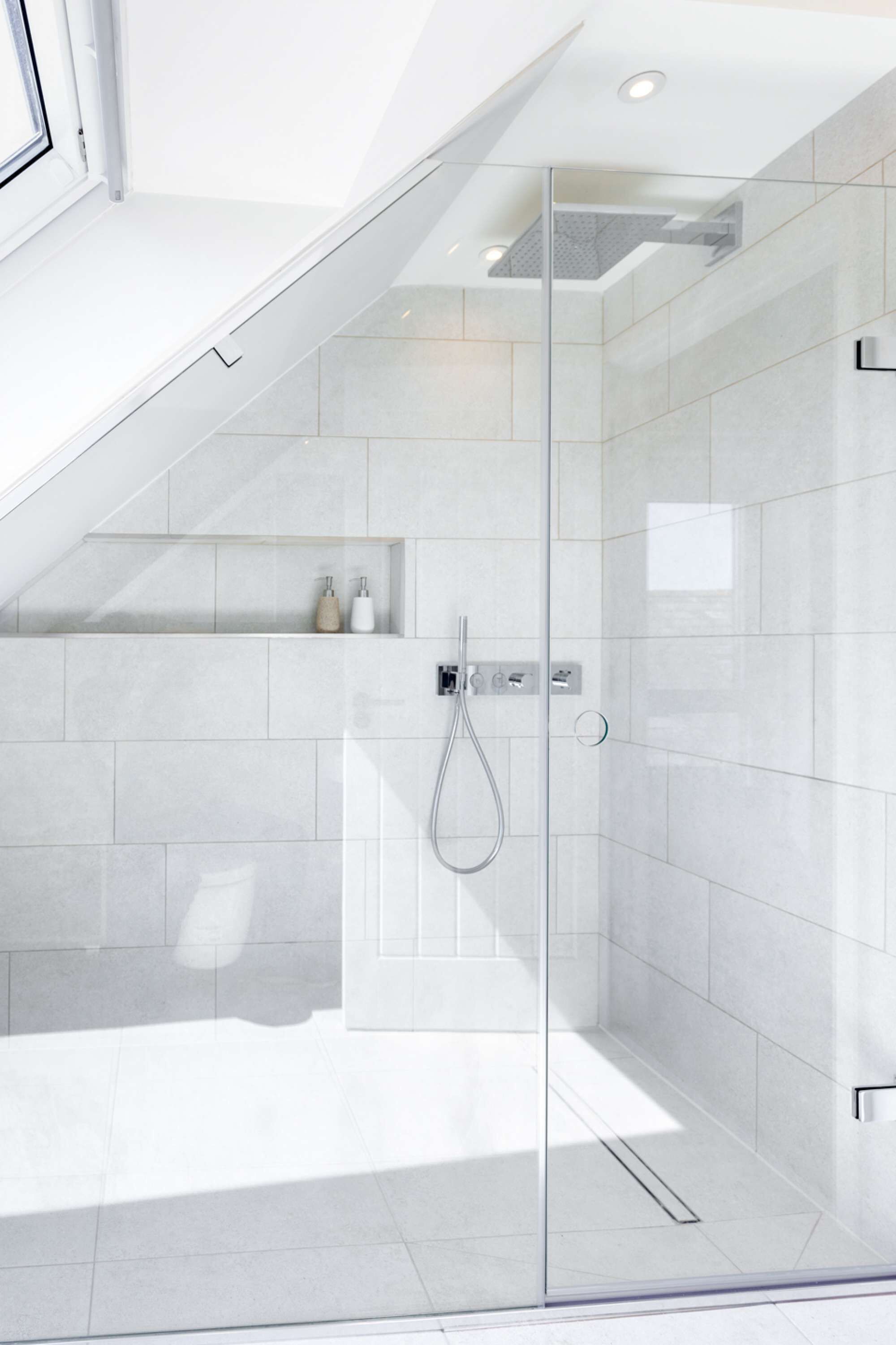 A modern shower with a glass door, white tiled walls, and a built-in shelf with toiletries.